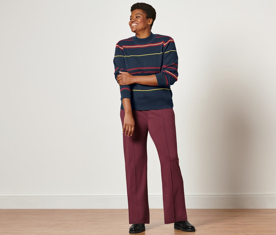 Eine Frau steht und trägt einen mehrfarbigen Feinstrickpullover mit Zopfmuster, Joggpants mit Biese und Lederboots.