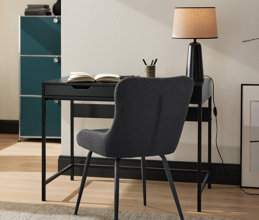 Ein anthrazitfarbener, gepolsterter Stuhl steht an einem schwarzen Schreibtisch. Auf dem Tisch stehen eine schwarze Tischleuchte mit Textilschirm und ein offenes Buch.