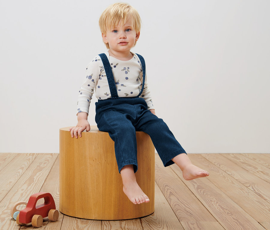Ein Junge sitzt auf einem Holzschemel und trägt eine Baby-Twillhose mit Latz.