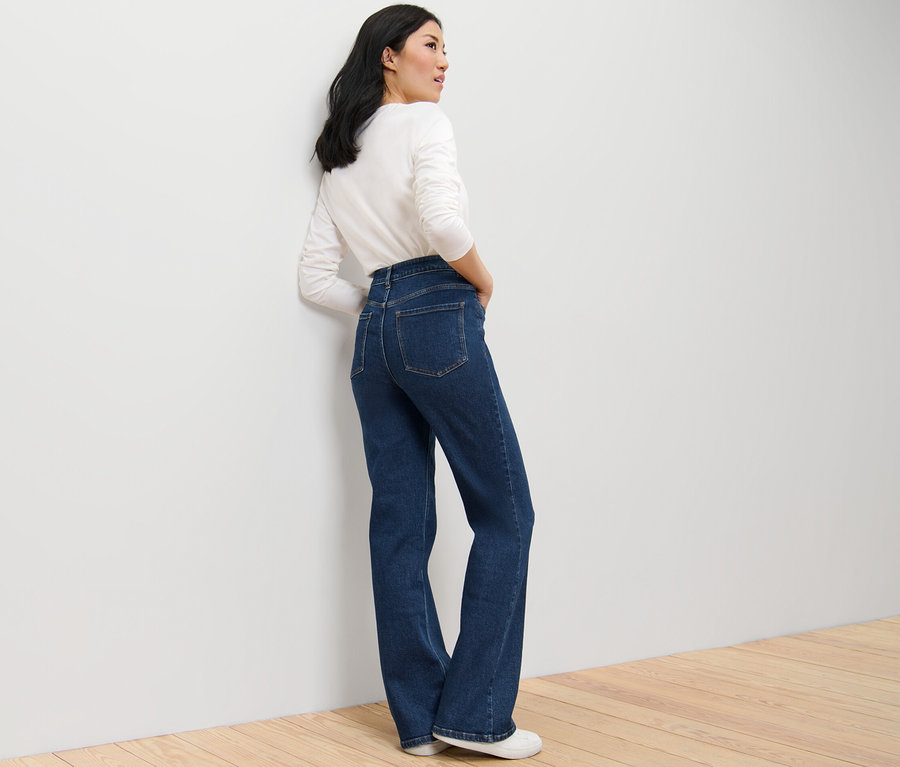 Eine Frau posiert in dunkelblauen Bootcut Jeans – Fit »Lou« und einem weißen Langarmshirt.