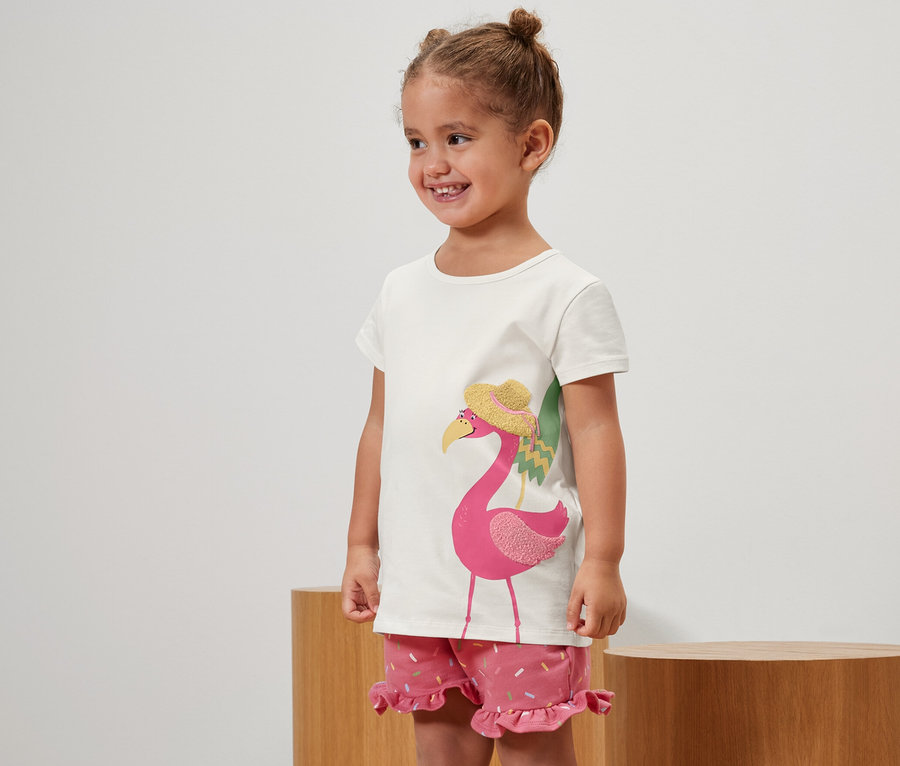 Lächelndes kleines Mädchen posiert in 2 Kinder-T-Shirts mit Applikation mit Flamingo und rosa Shorts.