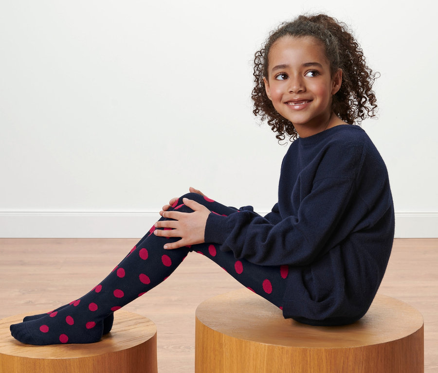 Mädchen sitzt auf einem Holzhocker und trägt eine dunkelblaue Strumpfhose mit roten Punkten, 2 Kleinkind-Strumpfhosen.