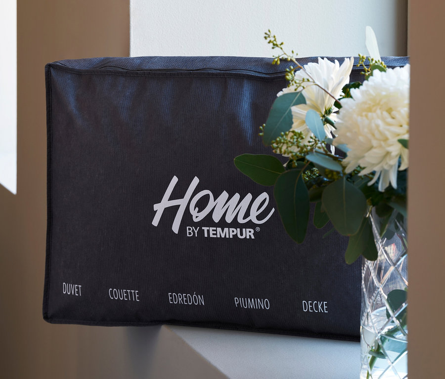 Eine verpackte Home By TEMPUR® kuschelige Luxe Duo-Decke neben einer Vase mit weißen Blumen.