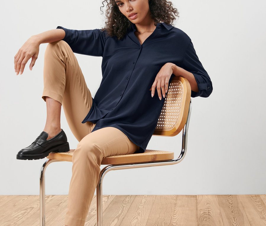 Frau sitzt auf einem Stuhl und trägt eine dunkelblaue Piqué-Jerseybluse und eine beige Hose.