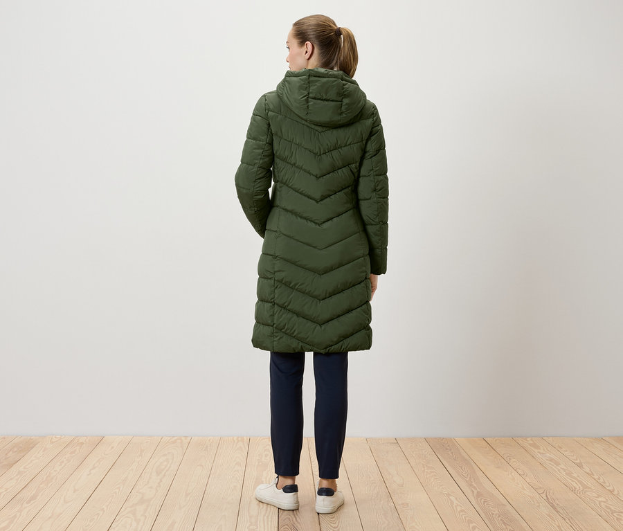 Eine Frau steht mit dem Rücken zur Kamera und trägt einen langen, grünen Steppmantel mit Kapuze, dunkelblaue Hosen und weiße Turnschuhe.