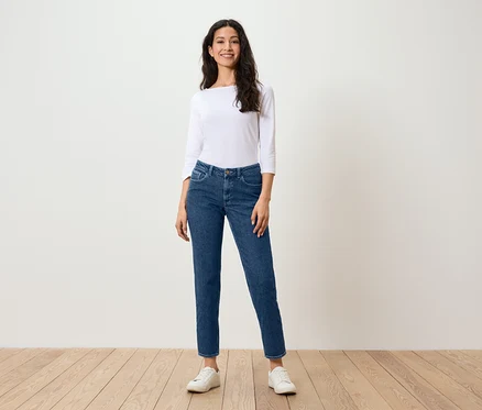 Eine Frau steht auf einem Holzboden und trägt blaue Jeans, ein weißes Hemd und weiße Turnschuhe.
