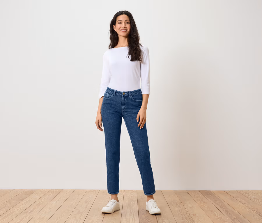 Eine Frau steht auf einem Holzboden und trägt blaue Jeans, ein weißes Hemd und weiße Turnschuhe.