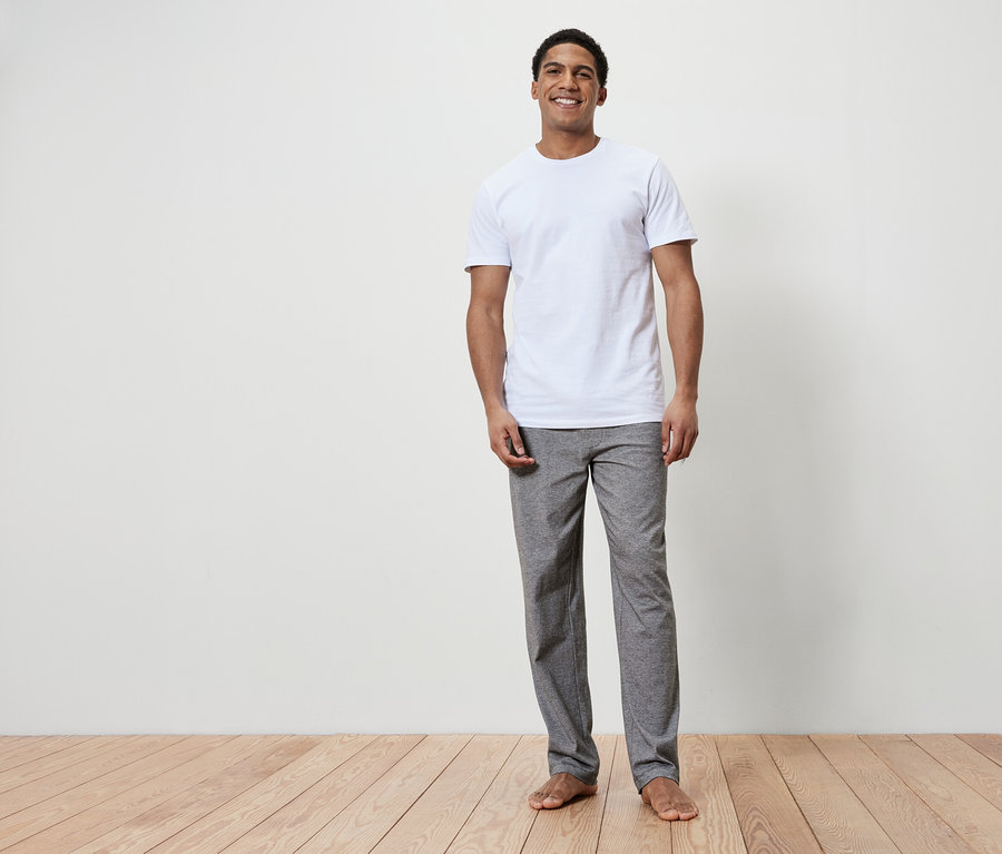 Ein lächelnder Mann steht barfuß auf einem Holzboden. Er trägt ein weißes T-Shirt und graue Hosen.