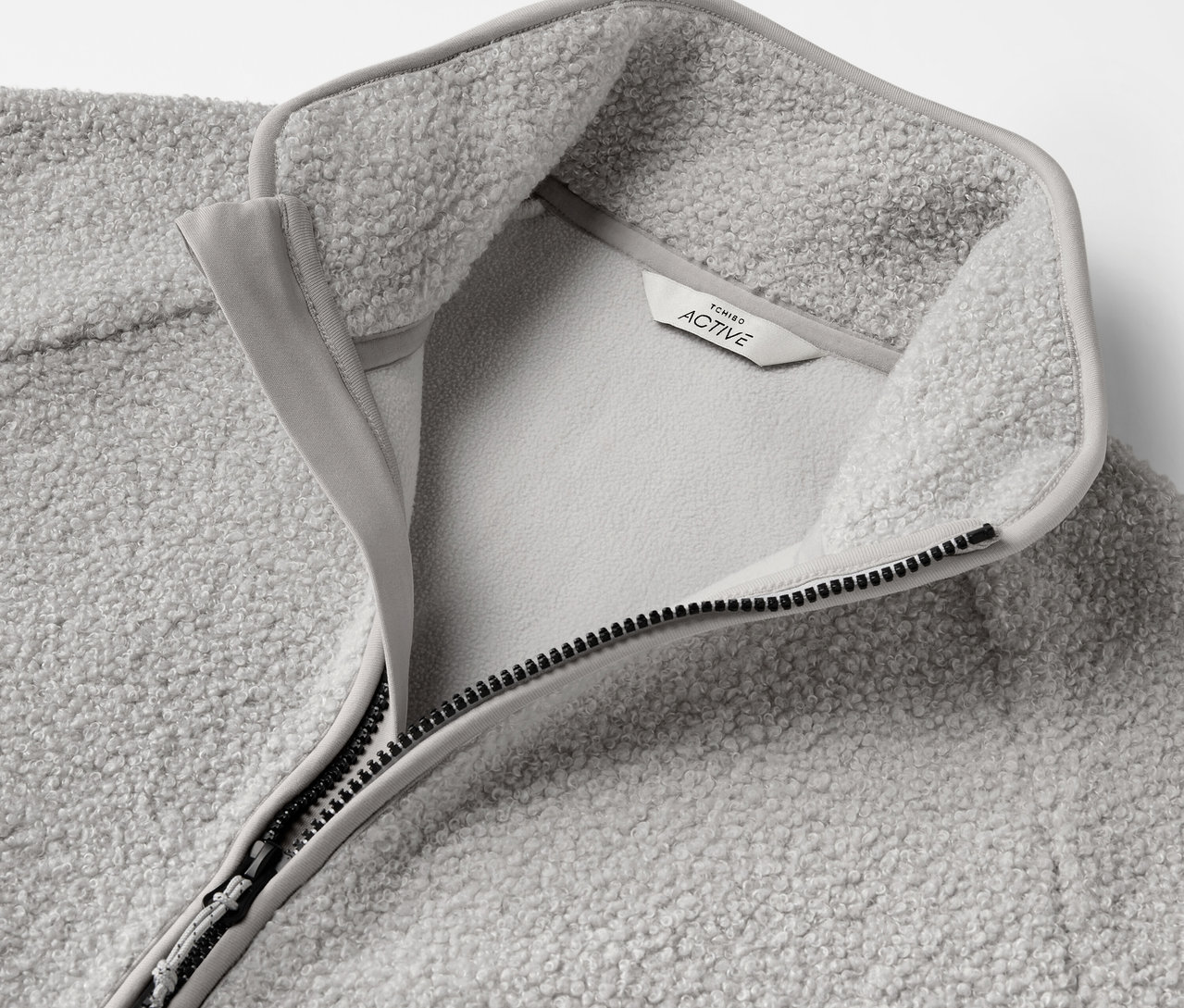 Detailaufnahme einer grauen Fleecejacke mit geöffnetem Reißverschluss und Etikett.