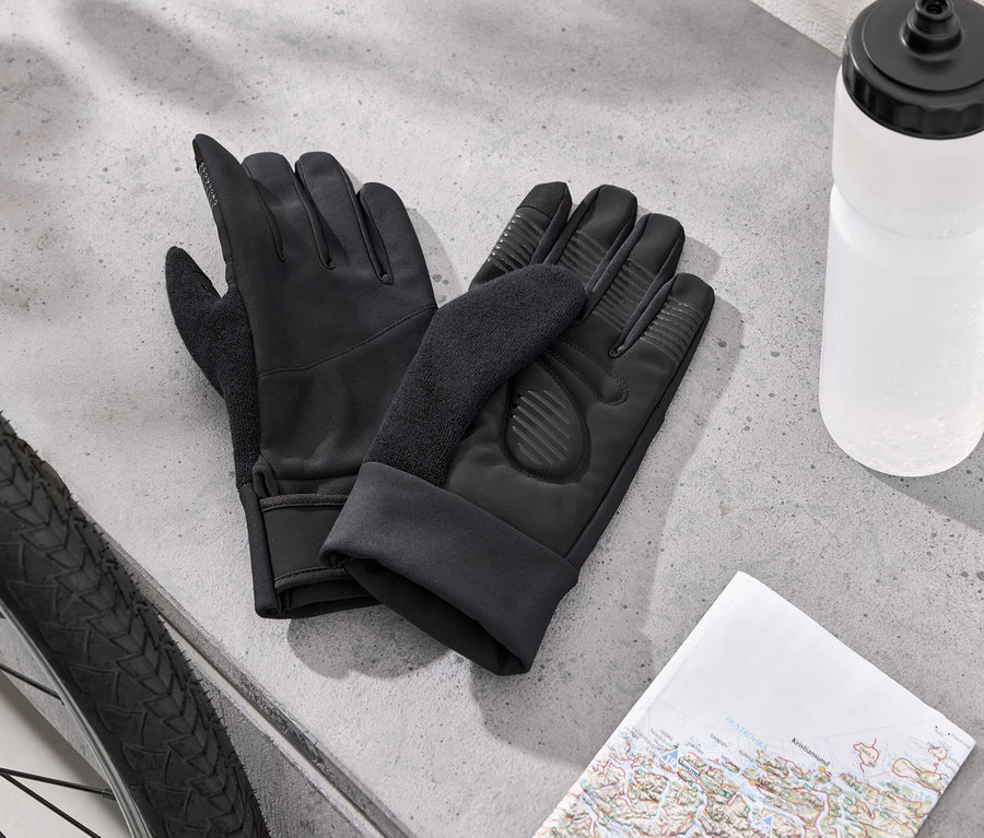 Zwei schwarze Fahrradhandschuhe liegen neben einem Fahrradreifen, einer weißen Wasserflasche und einer Karte auf einer grauen Oberfläche.