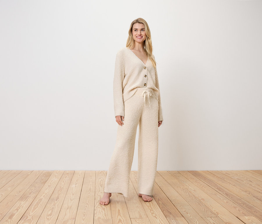 Blonde Frau steht in einem cremefarbenen Cardigan und Hose auf einem Holzboden.