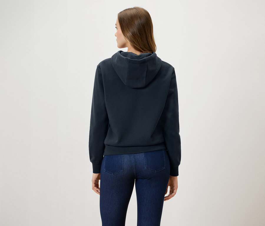 Frau steht mit dem Rücken zum Betrachter und trägt einen dunkelblauen Kapuzenpullover und Jeans.