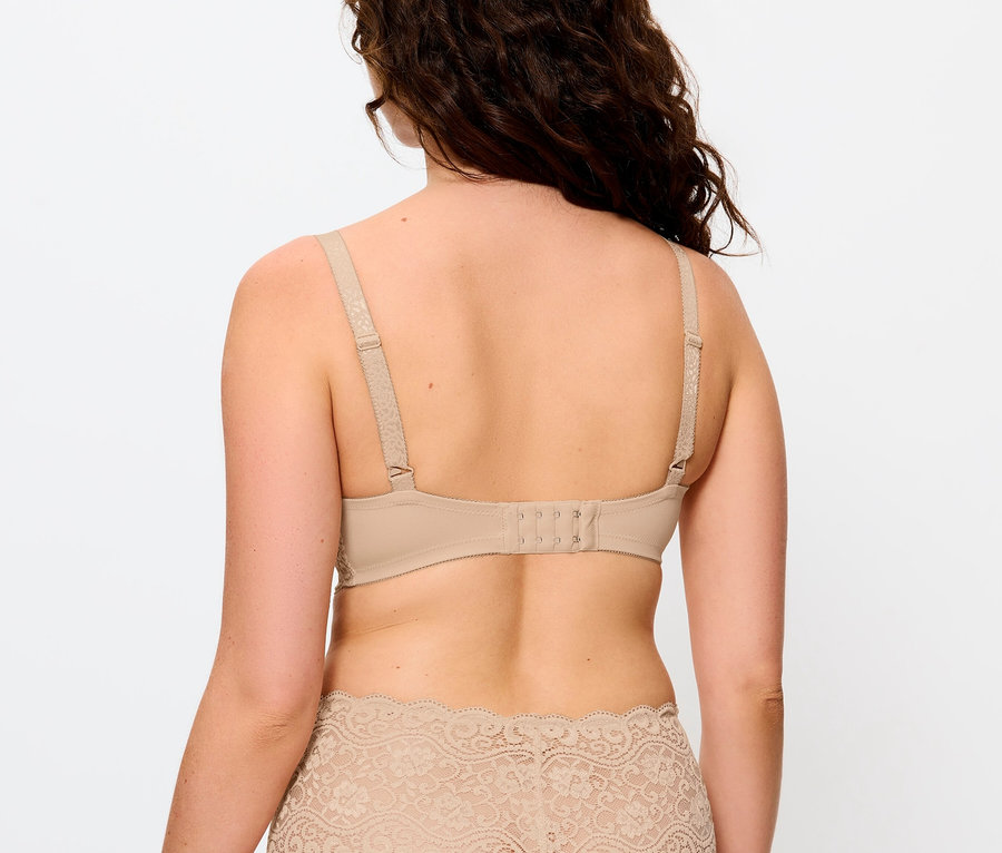 Rückansicht einer Frau mit braunem, gewelltem Haar, die einen beigen BH und Höschen mit Spitze trägt.