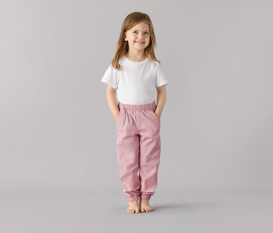 Ein lächelndes Mädchen steht mit den Händen in den Taschen einer Kinder-Pull on pants ‒ Fit »Ella«, altrosa.