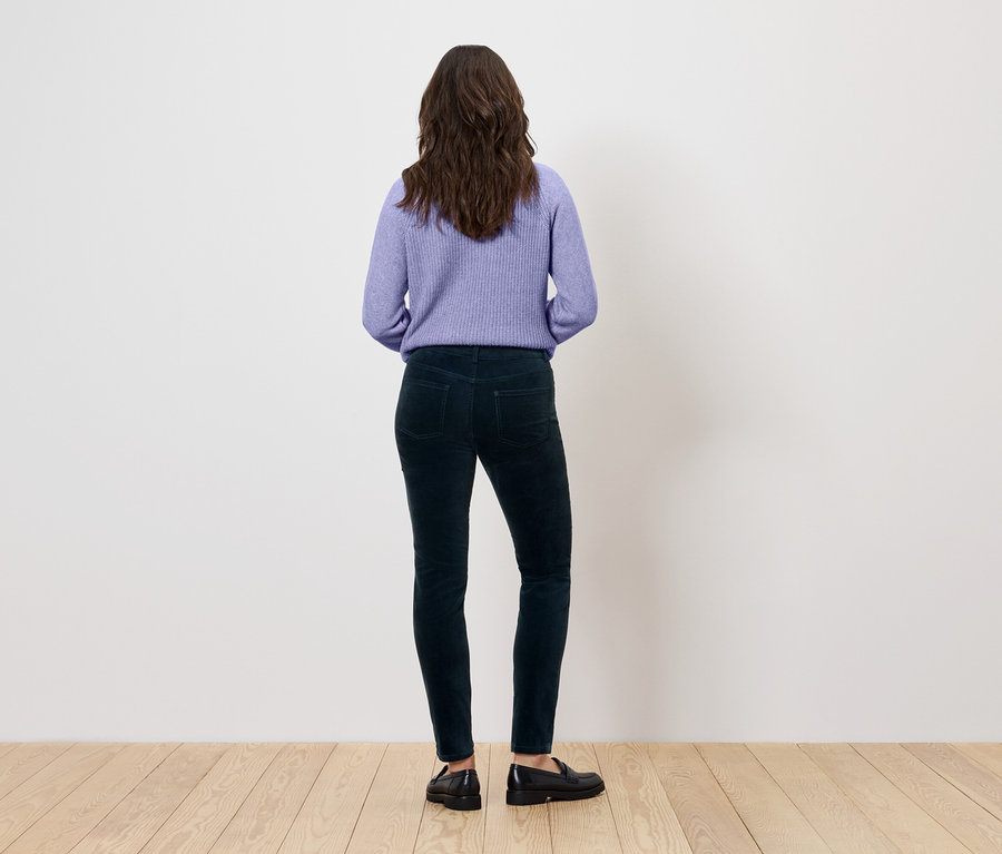Rückansicht einer Frau mit braunem, gewelltem Haar, die einen lila Pullover, dunkelgrüne Hosen und schwarze Schuhe trägt.