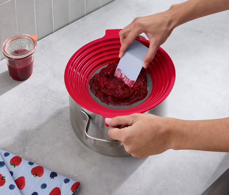 Eine Person passiert rote Früchte mit einem 2-in-1: Mehl-und-Passiersieb in einen Topf. Daneben steht ein Glas Marmelade und ein Geschirrtuch mit Erdbeermuster.