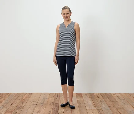 Eine Frau steht vor einer weißen Wand und trägt ein blau-weiß gestreiftes Tanktop, blaue Caprihosen und Ballerinaschuhe.