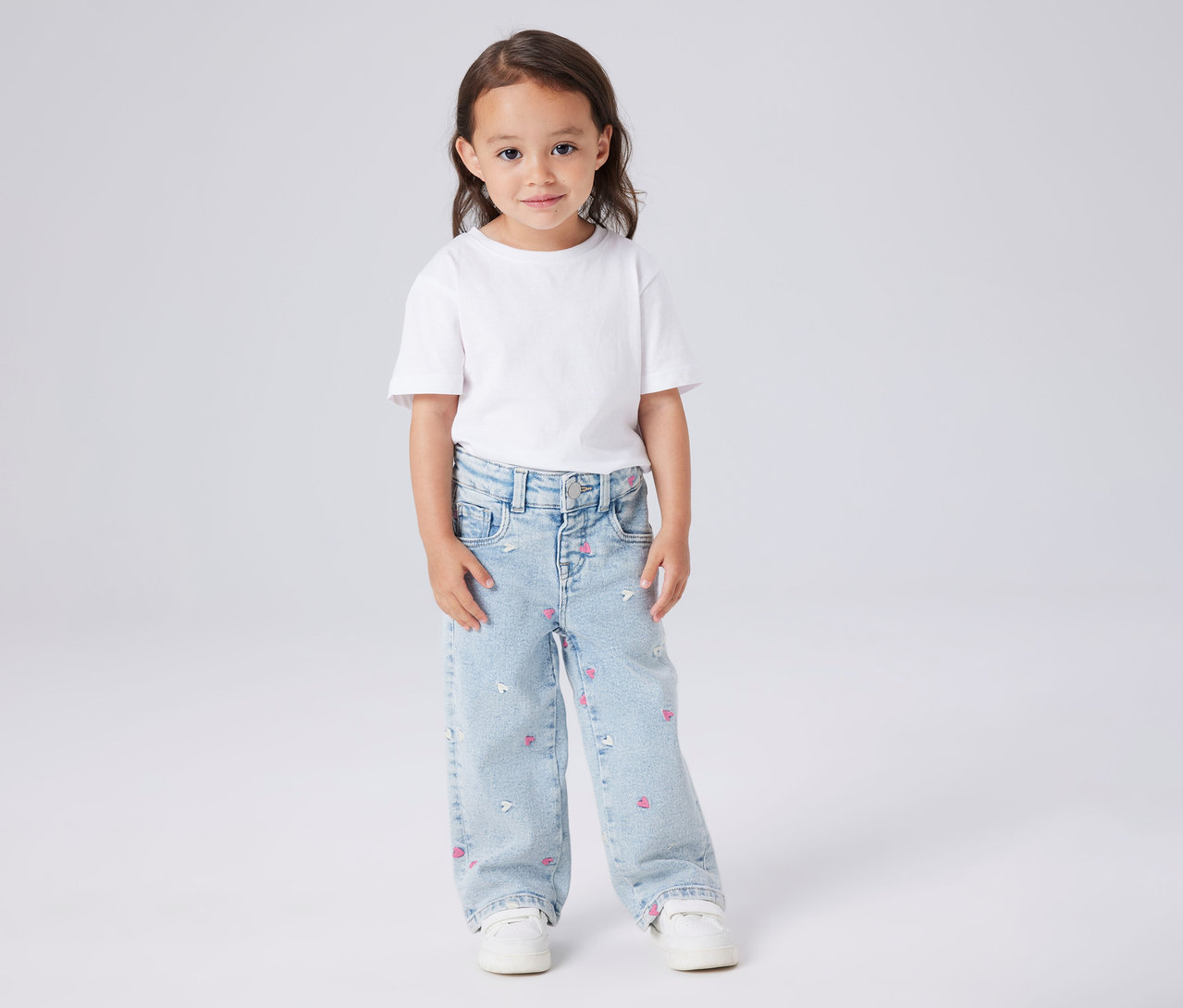 Mädchen trägt ein weißes T-Shirt und NAME IT Kinder-Wide Leg Jeans mit rosa Herzchen und weißen Maus-Motiven.