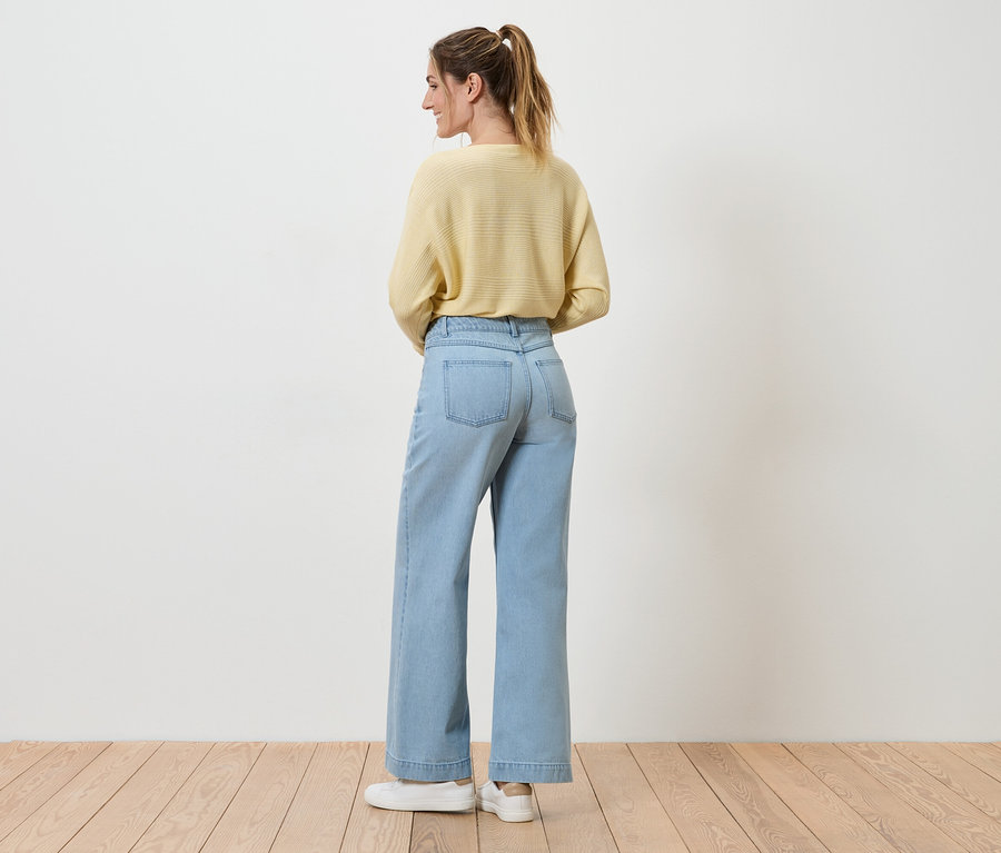 Frau mit Pferdeschwanz trägt hellgelben Pullover, weite, hellblaue Jeans und weiße Sneaker, von hinten gesehen.