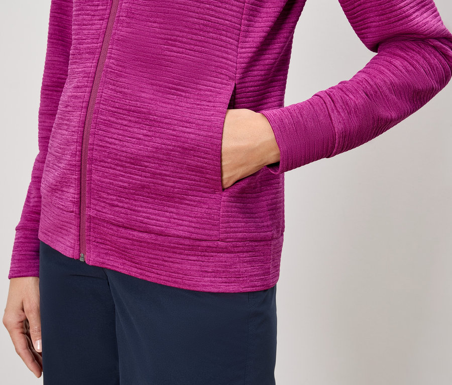 Nahaufnahme einer Person, die eine pinkfarbene Jacke mit Reißverschluss und marineblaue Hosen trägt und eine Hand in die Jackentasche steckt.