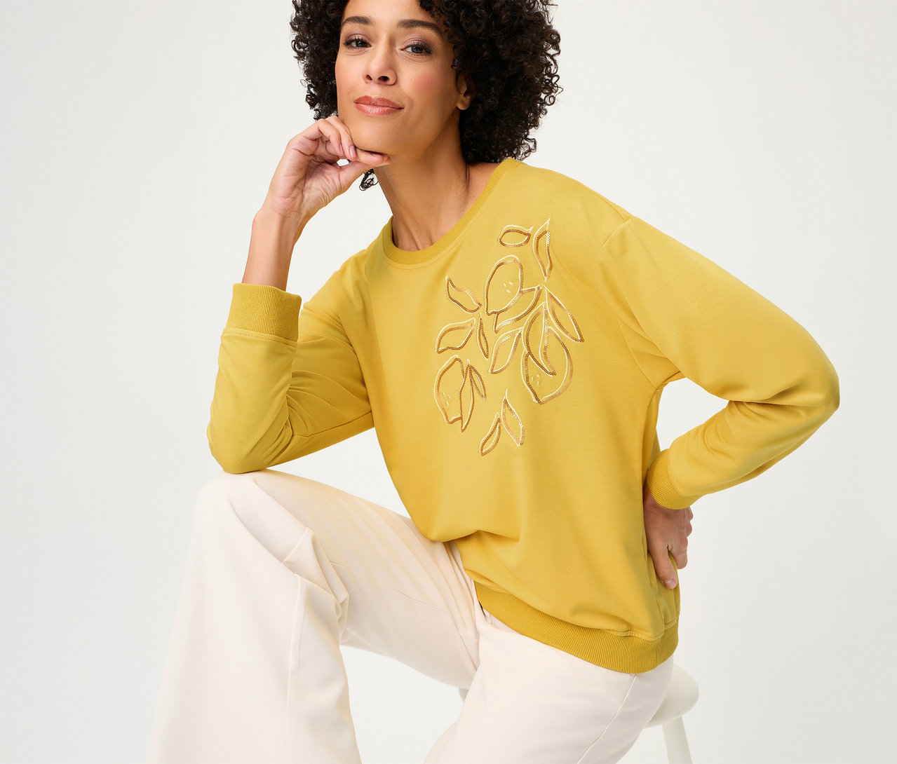 Frau sitzt auf einem Hocker und trägt ein Olsen Sweatshirt Long Sleeves in Gelb mit Zitronen-Applikation und eine weite, helle Hose.