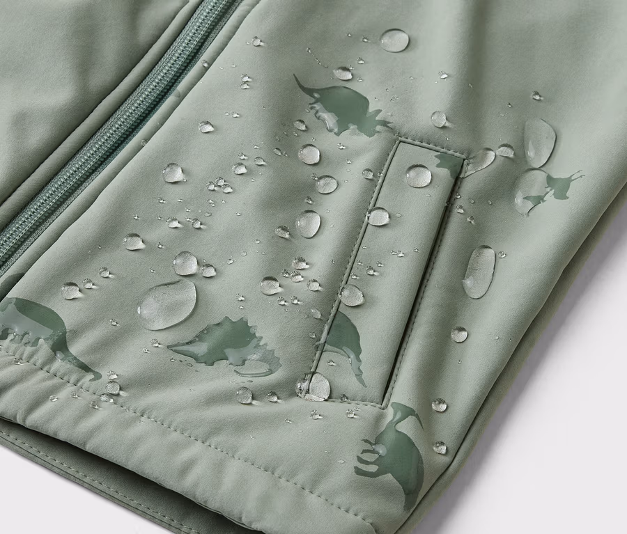 Nahaufnahme einer hellgrünen Jacke mit Reißverschluss, Tasche und Dinosaurier-Muster, bedeckt mit Wassertropfen.