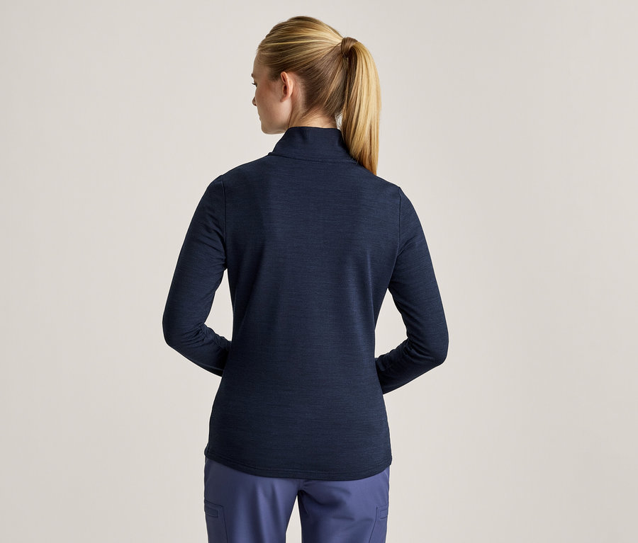Frau von hinten, mit blondem Pferdeschwanz, trägt ein dunkelblaues Langarmshirt und blaue Hose.