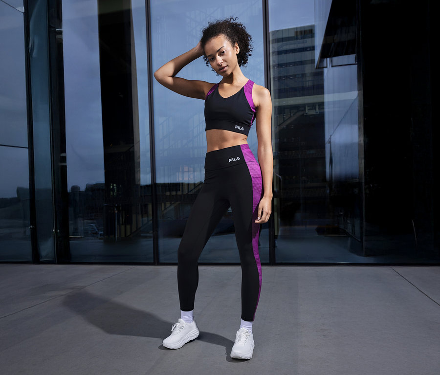 Frau in schwarzem Sport-BH und Leggings mit lila Streifen posiert vor einem Gebäude.
