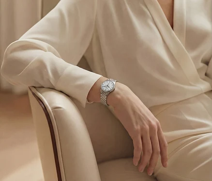 Eine Frau in einem weißen Hosenanzug sitzt auf einem Stuhl und trägt eine silberne Armbanduhr.