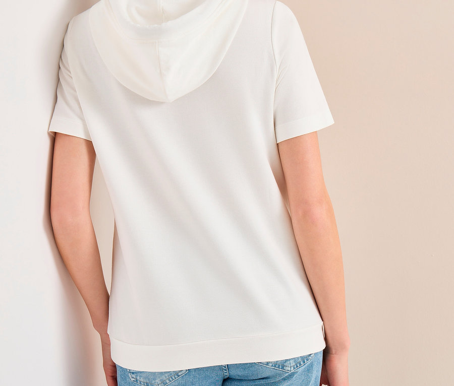 Frau von hinten, an eine Wand gelehnt und nach rechts blickend, trägt ein weißes kurzärmeliges CECIL Hoodie-Shirt und blaue Jeans.