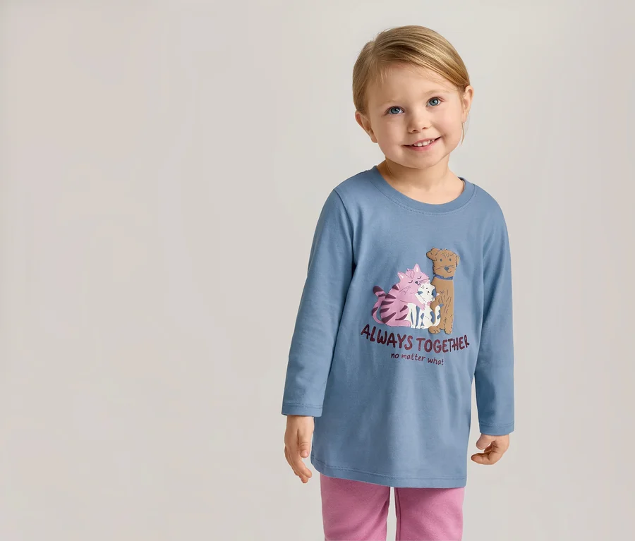 Lächelndes Mädchen mit blauem Langarmshirt mit Katzen- und Hundemotiv und dem Schriftzug "Always Together" und rosafarbener Hose.
