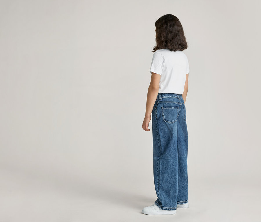 Rückansicht einer Person mit weißem T-Shirt, blauen Jeans und weißen Schuhen vor hellem Hintergrund.