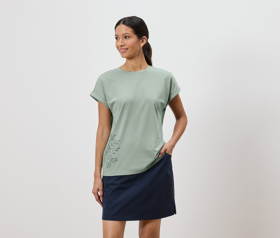 Frau mit hellgrünem T-Shirt mit Blumenmuster und blauem Rock, eine Hand in der Rocktasche.