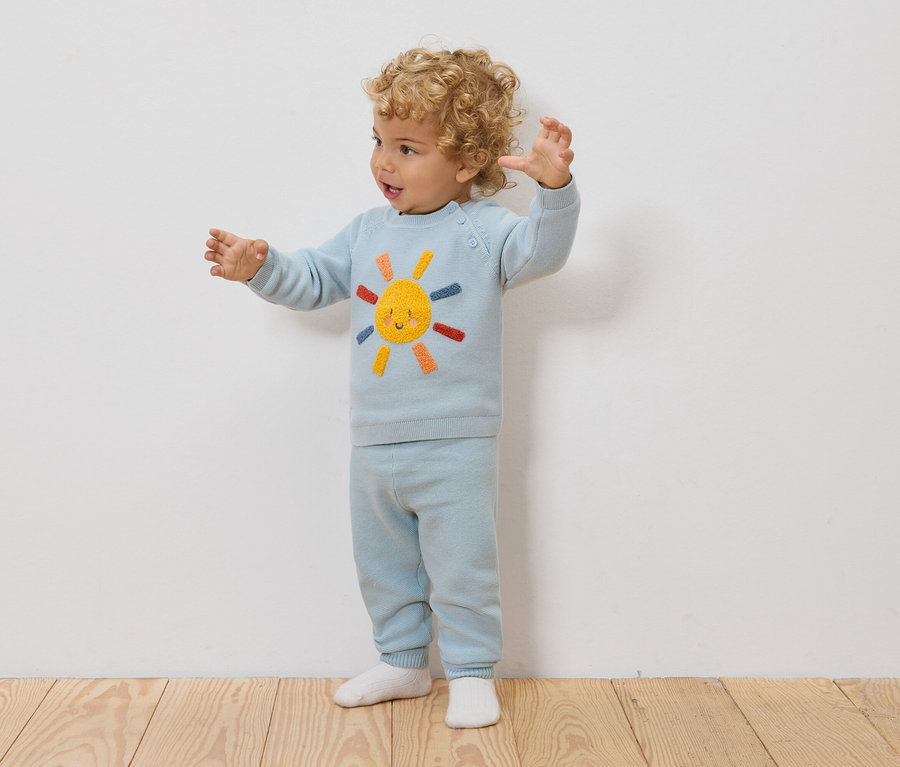 Ein Kleinkind mit lockigen Haaren steht vor einer weißen Wand und trägt einen hellblauen Pullover und eine Hose mit einem Sonnenmotiv.
