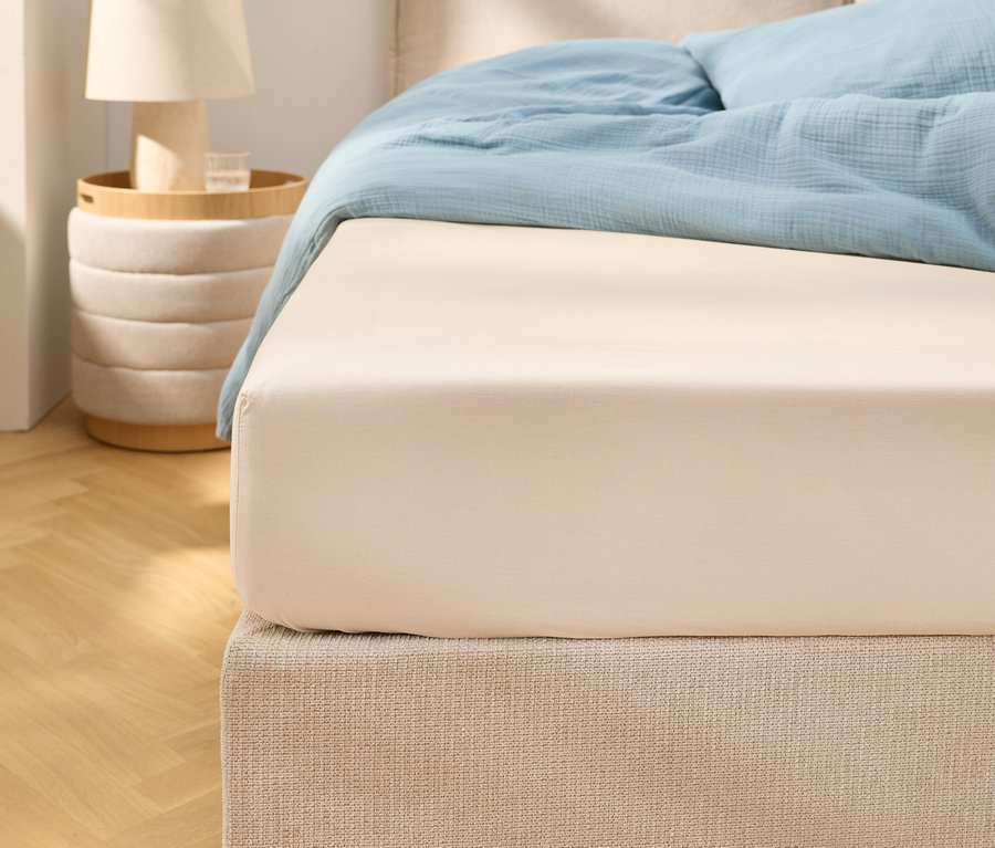 Nahaufnahme eines beigen Spannbettlakens auf einem Bett mit hellblauer Bettwäsche. Ein Nachttisch mit einer Lampe steht daneben.