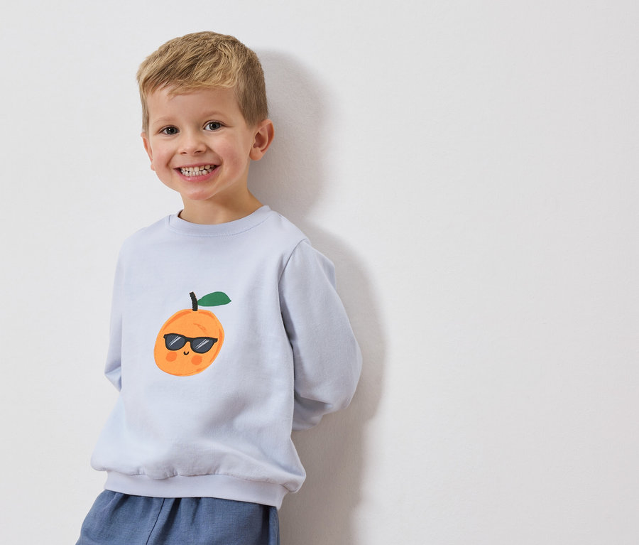 Lächelnder Junge trägt hellblaues Sweatshirt mit einer Orange mit Sonnenbrille darauf.