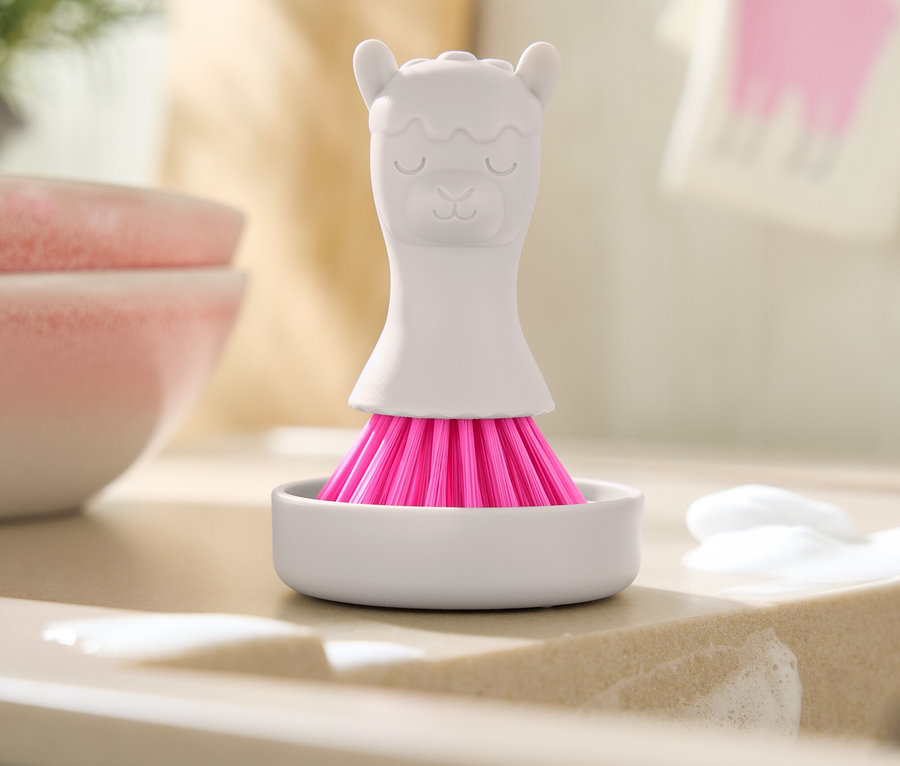 Eine weiße Bürste mit rosa Borsten in Form eines Lamas steht in einer weißen Schale auf einer Spüle mit Seifenresten.