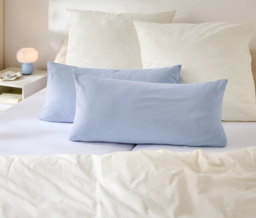 Auf einem Bett liegen zwei blaue und zwei weiße Kopfkissen. Auf dem Nachttisch steht eine Lampe.