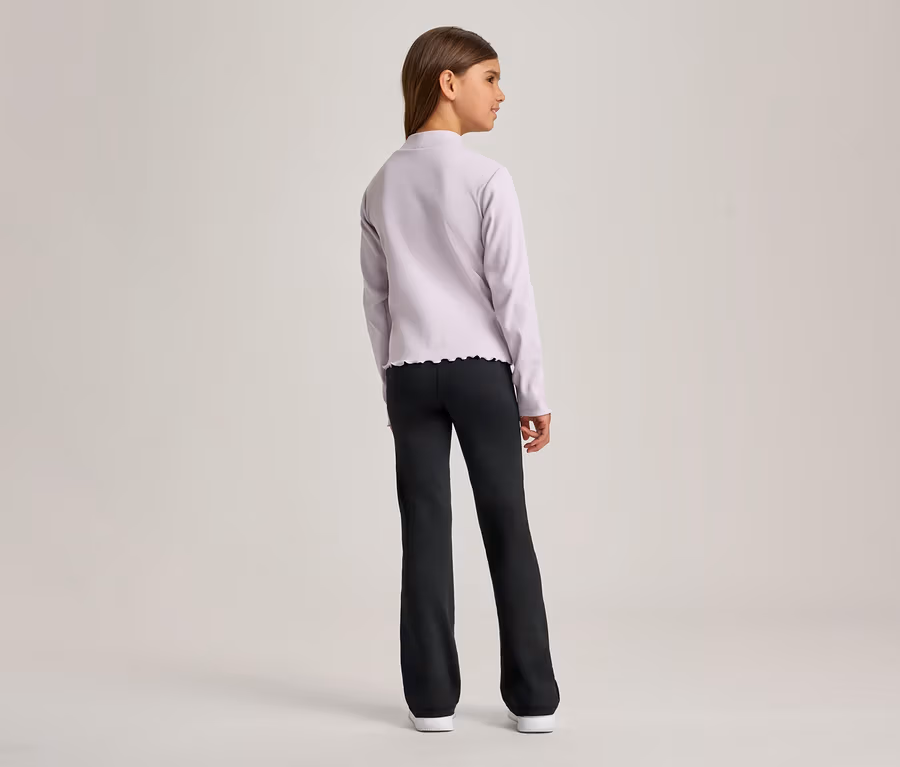 Ein junges Mädchen steht mit dem Rücken zum Betrachter und blickt nach rechts. Sie trägt ein hellviolettes Langarmshirt mit Rüschensaum und eine schwarze ausgestellte Hose, dazu weiße Schuhe.