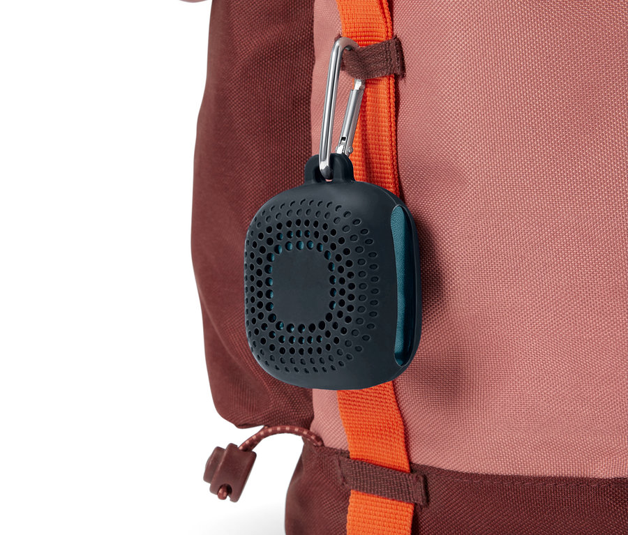 Dunkelgrauer, gelochter Behälter mit Karabinerhaken hängt an einem orangefarbenen Riemen eines rotbraunen Rucksacks.