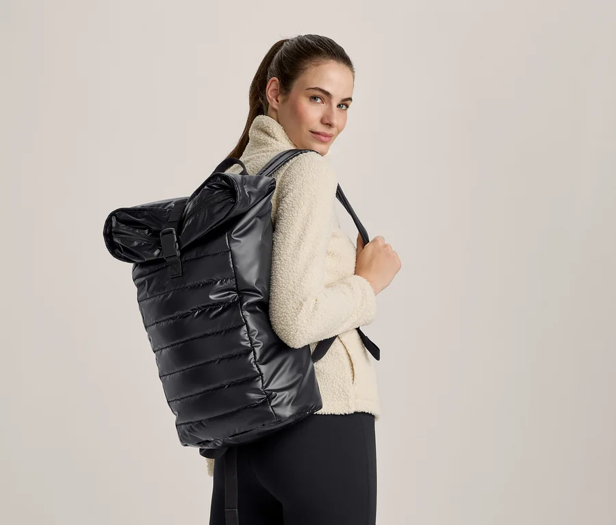 Frau mit glänzendem schwarzem Stepp-Roll-Top-Rucksack und cremefarbener Fleecejacke, die über die Schulter blickt.