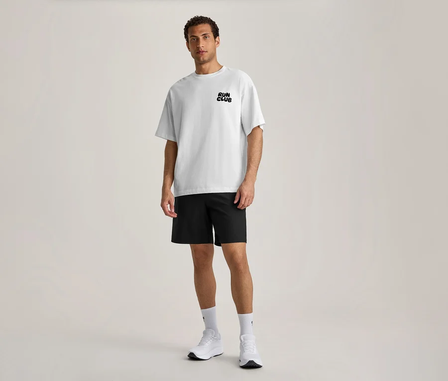 Ein Mann steht nach vorne blickend und trägt ein weißes Sportshirt mit dem schwarzen Schriftzug „RUN CLUB“ auf der Brust, schwarze Shorts, weiße Socken und weiße Sportschuhe.