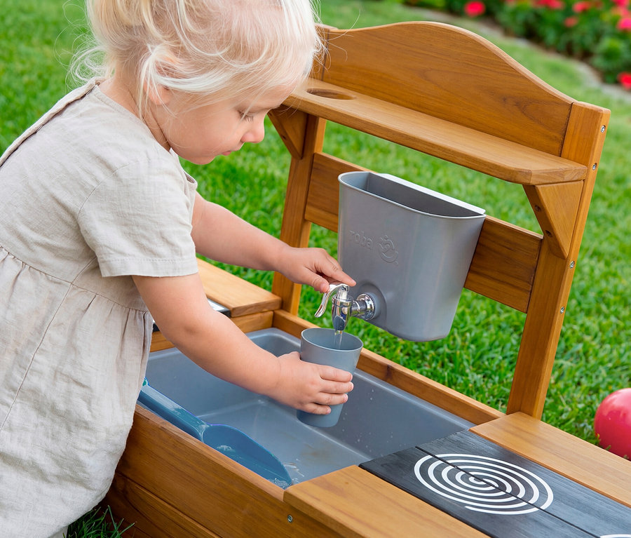 Kleines blondes Kind füllt einen grauen Becher mit Wasser aus einem silbernen Wasserhahn an einer hölzernen Outdoor-Spielküche, mit grauem Becken und Herdplatte.