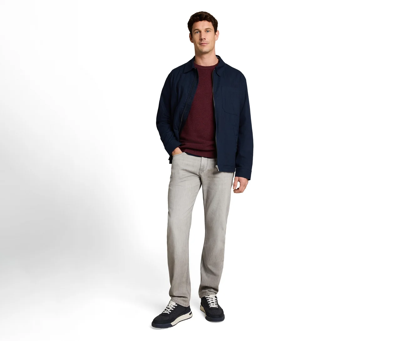 Stehender Mann in dunkelblauer Jacke, bordeauxrotem Pullover, hellgrauer Jeans und dunkelblauen Sneakern. Seine linke Hand steckt in der Hosentasche.
