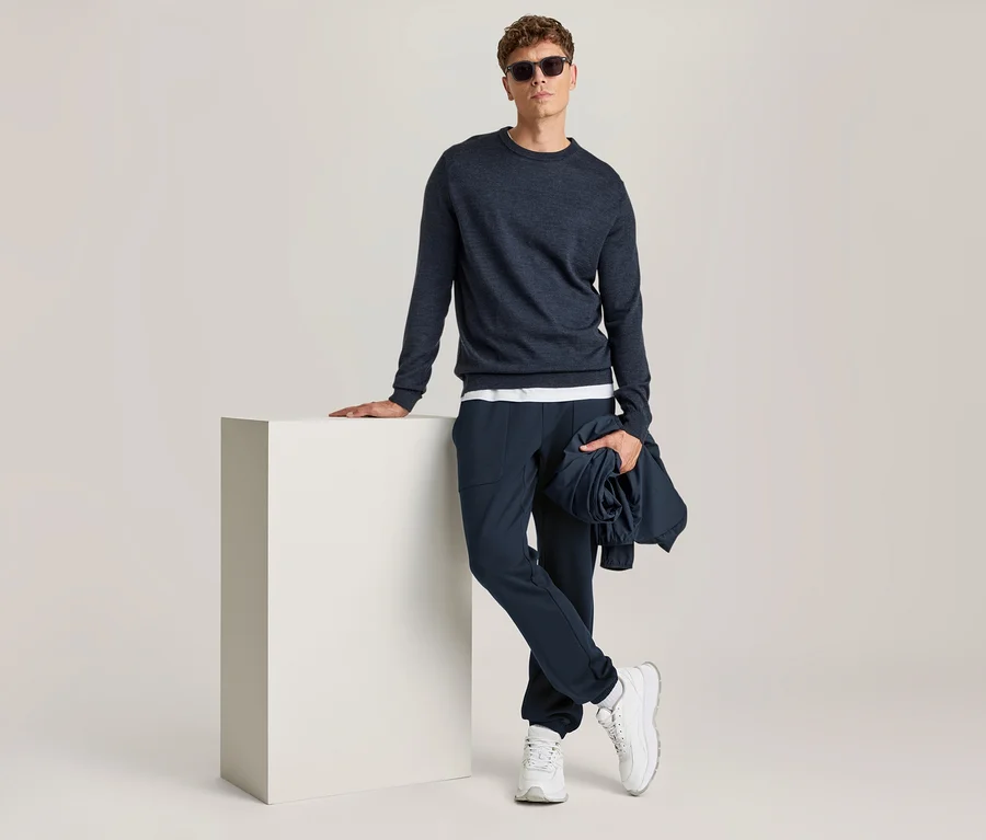Mann steht, lehnt an einem weißen Würfel, trägt dunkelblaues Langarmshirt, dunkelblaue Hose, weiße Sneaker und Sonnenbrille, hält eine zusammengerollte dunkelblaue Jacke.