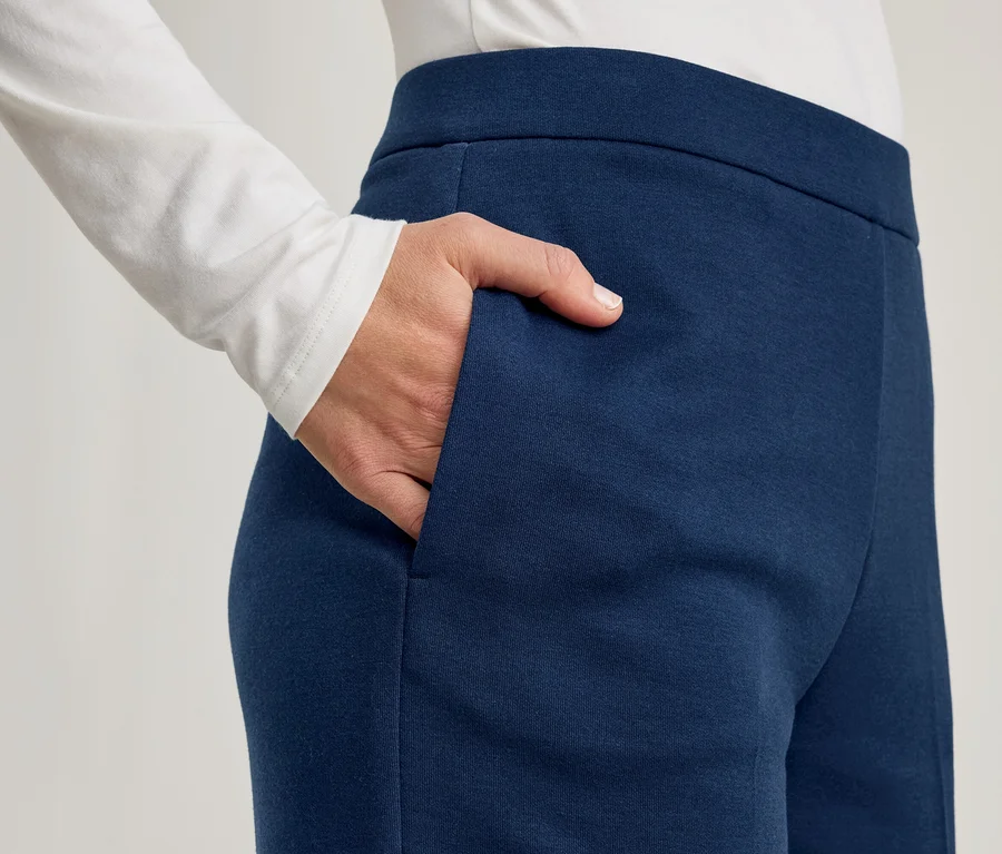 Eine Person trägt ein weißes Langarmshirt und eine dunkelblaue Hose, wobei die rechte Hand in der Hosentasche steckt.