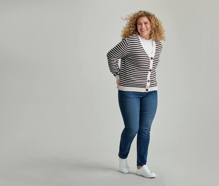 Lächelnde Frau mit lockigem Haar in gestreifter Strickjacke, weißem T-Shirt, blauen Jeans und weißen Turnschuhen, die Hände hinter dem Rücken haltend.