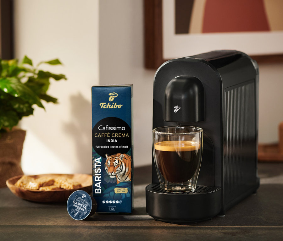 Schwarze Tchibo Cafissimo Kaffeemaschine mit einem Glas Kaffee, daneben eine Packung Tchibo Cafissimo Caffè Crema India Kaffeekapseln und eine einzelne Kapsel.