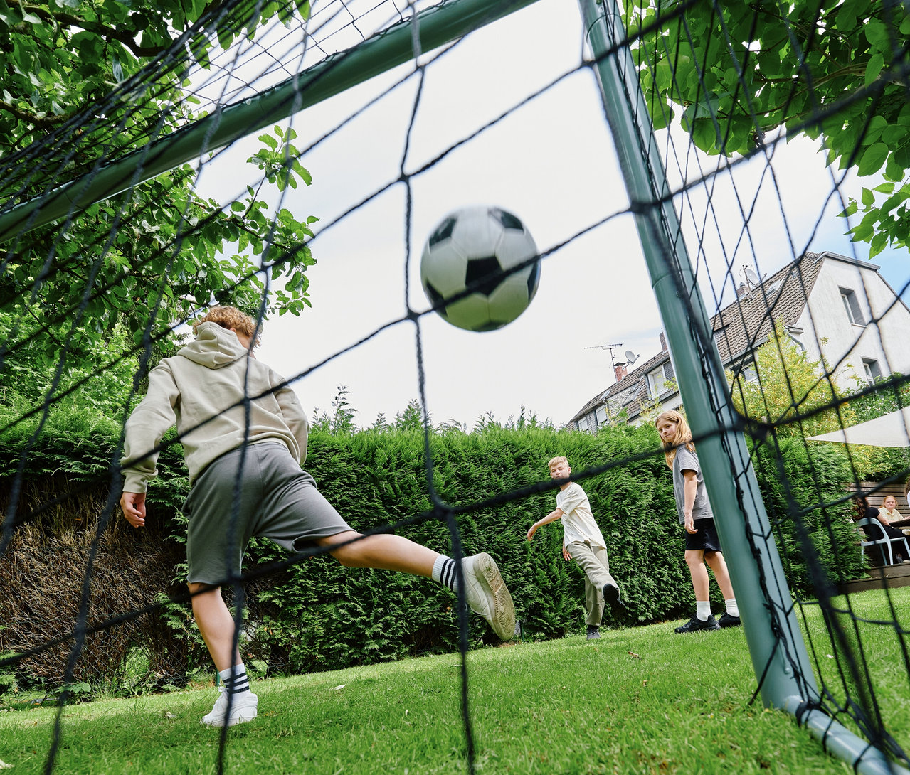 Blick durch das Netz eines HUDORA Fußballtors »Expert 300« auf ein Kind, das einen Fußball darauf schießt. Der Ball fliegt ins Tor, während zwei weitere Kinder im Garten zusehen.