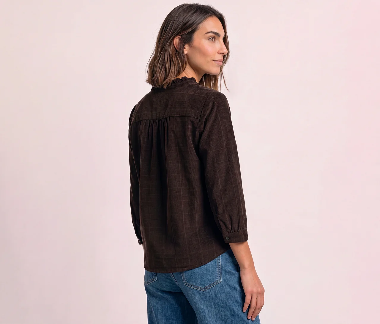 Frau trägt eine braune Street One Cordbluse mit Rüschenkragen und gerafftem Rücken sowie blaue Jeans, seitlich blickend vor hellrosa Hintergrund.